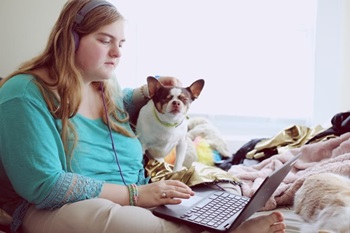 Woman with headphones and her dog sitting on a bed working on a computer