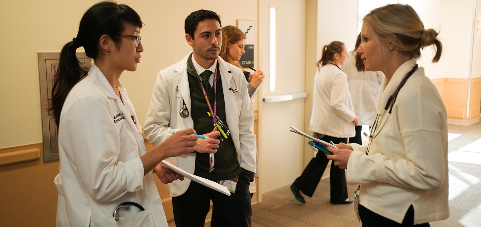 twostudentsandphysicianinahospitalhallwaytalkingandholdingclipboards