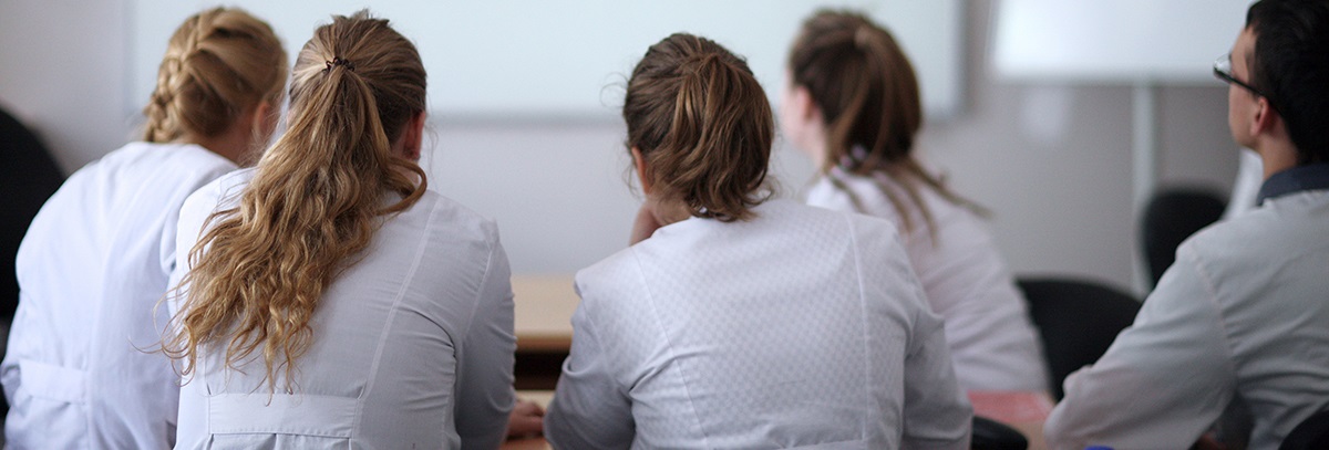 fivestudentsinwhitecoatsseenfrombackinclassroomandlookingatascreen