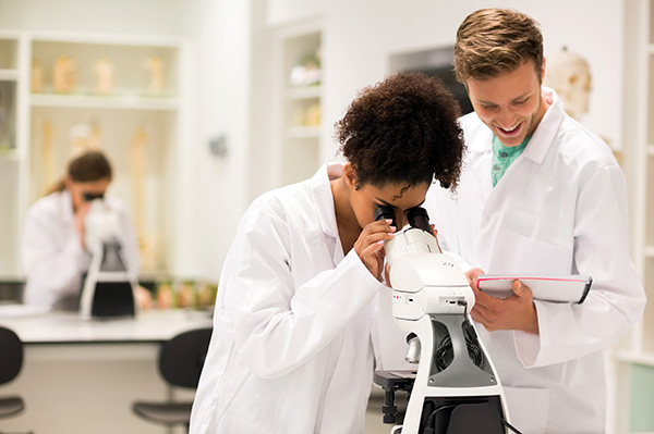 female student looking in microscope, male student standing near her and writing something in a notepad
