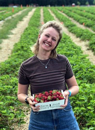 Ry Hemond standing on a farm, holding a box of berries. Wearing jeans and striped top.