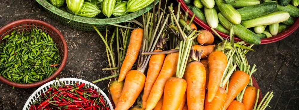 Assorted fresh vegetables, including carrots, green and red chili peppers, bitter melons, and cucumbers, displayed in baskets and on a surface.