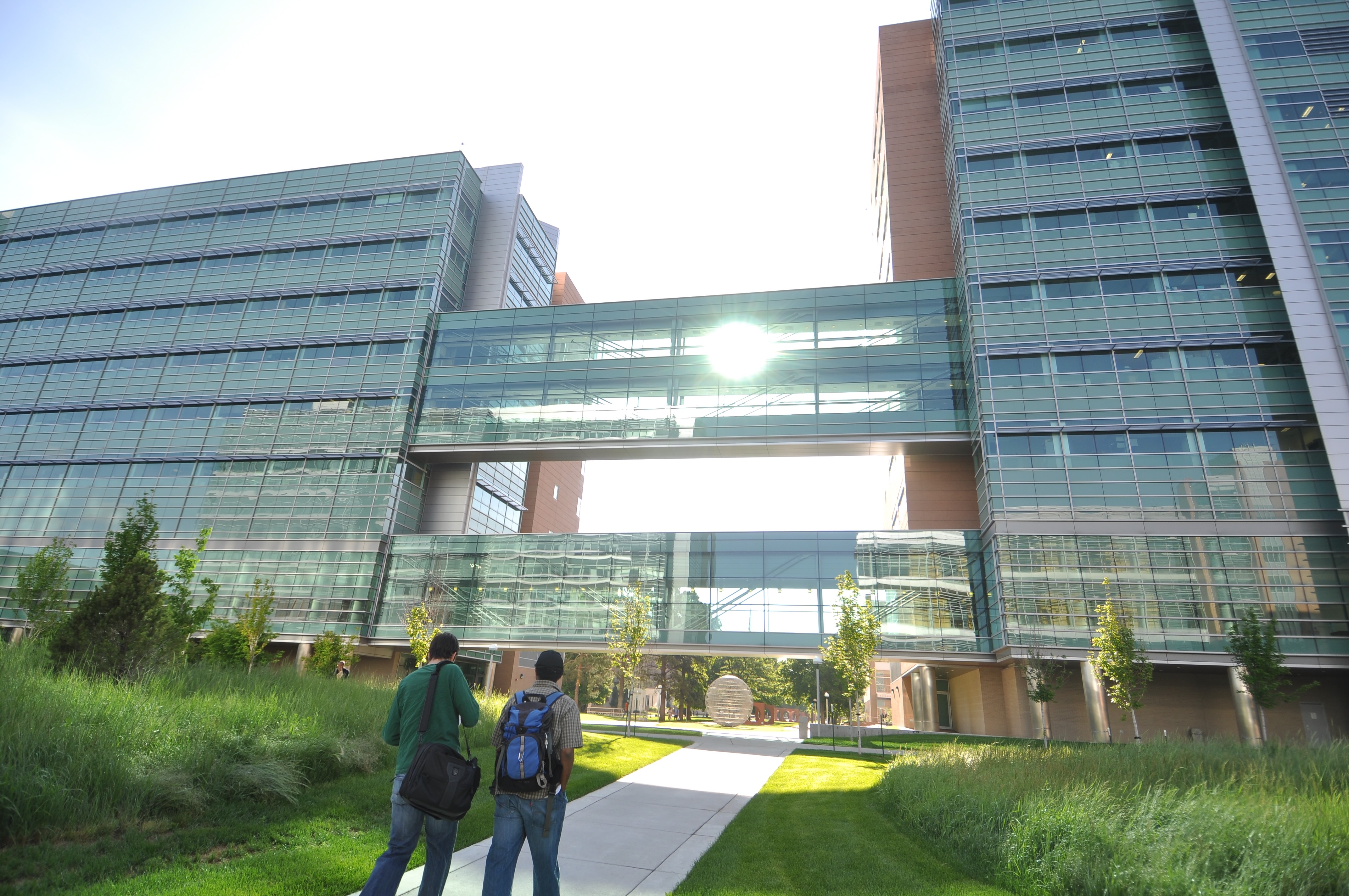 Two people walking toward the Research 1 North and South buildings, the sun peering through the glass walkways between the buildings