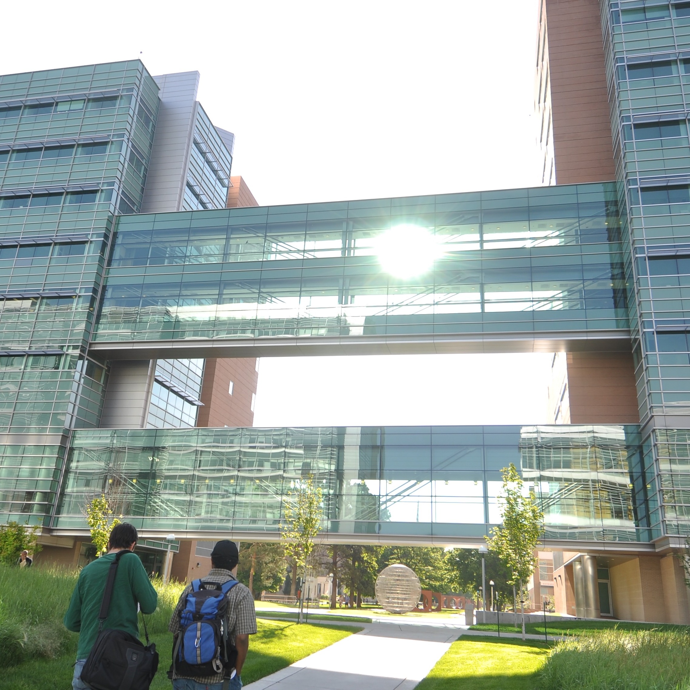 Two people walking toward the Research 1 North and South buildings, the sun peering through the glass walkways between the buildings