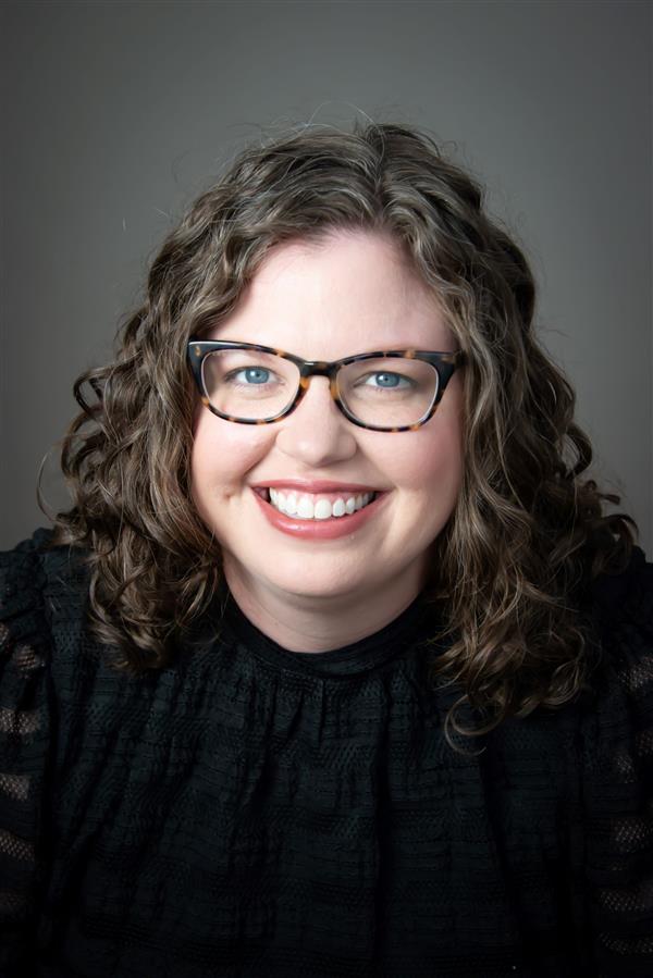 female with curly hair and brown and black glasses with a black shirt on smiling for a headshot