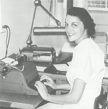 Black and white photo of Phyllis Horst, RN, seated at a desk in a white uniform, typing on a typewriter beside an old-fashioned lamp and office equipment.