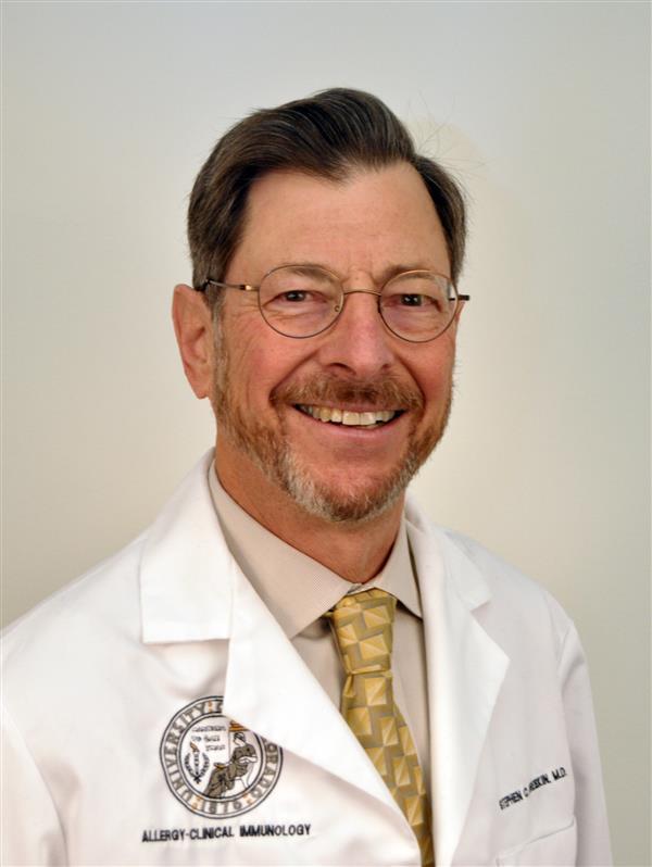 Professional headshot of a smiling man with glasses and a beard wearing a white medical coat and patterned tie against a light background.