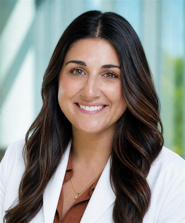 Professional headshot of a smiling woman with long dark brown hair wearing a white medical coat over a rust-colored blouse.