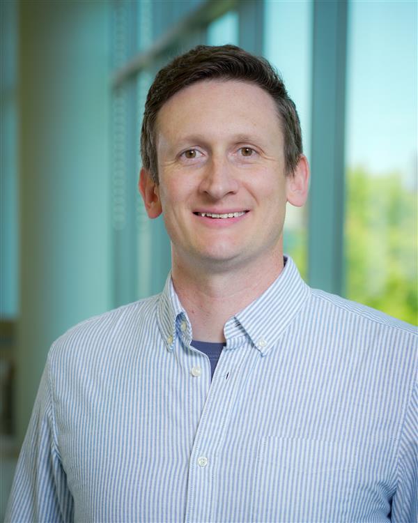 Professional headshot of a smiling man with short brown hair wearing a light blue striped button-down shirt in a bright indoor setting.