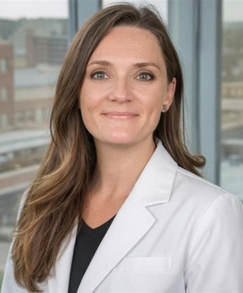 Professional headshot of a smiling woman with long light brown hair wearing a white medical coat over a black top, with windows and buildings in the background.