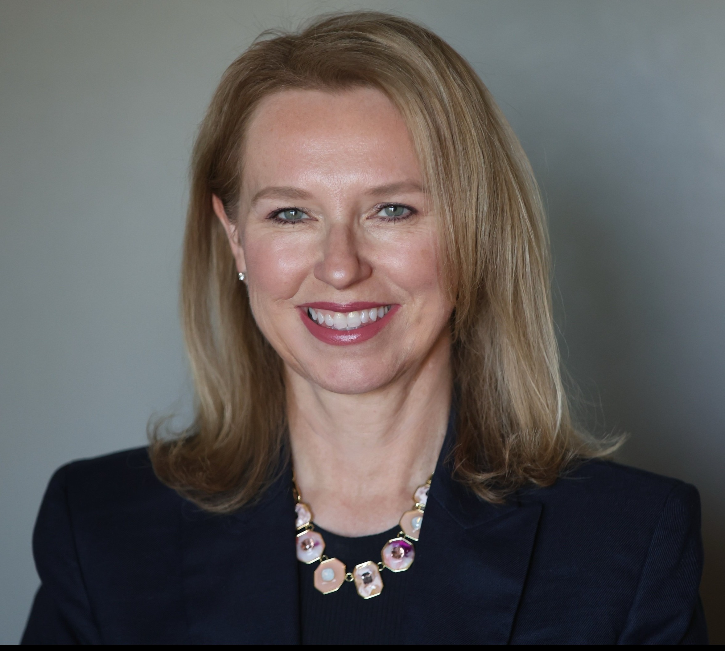 Headshot of woman in navy blazer with blank background.