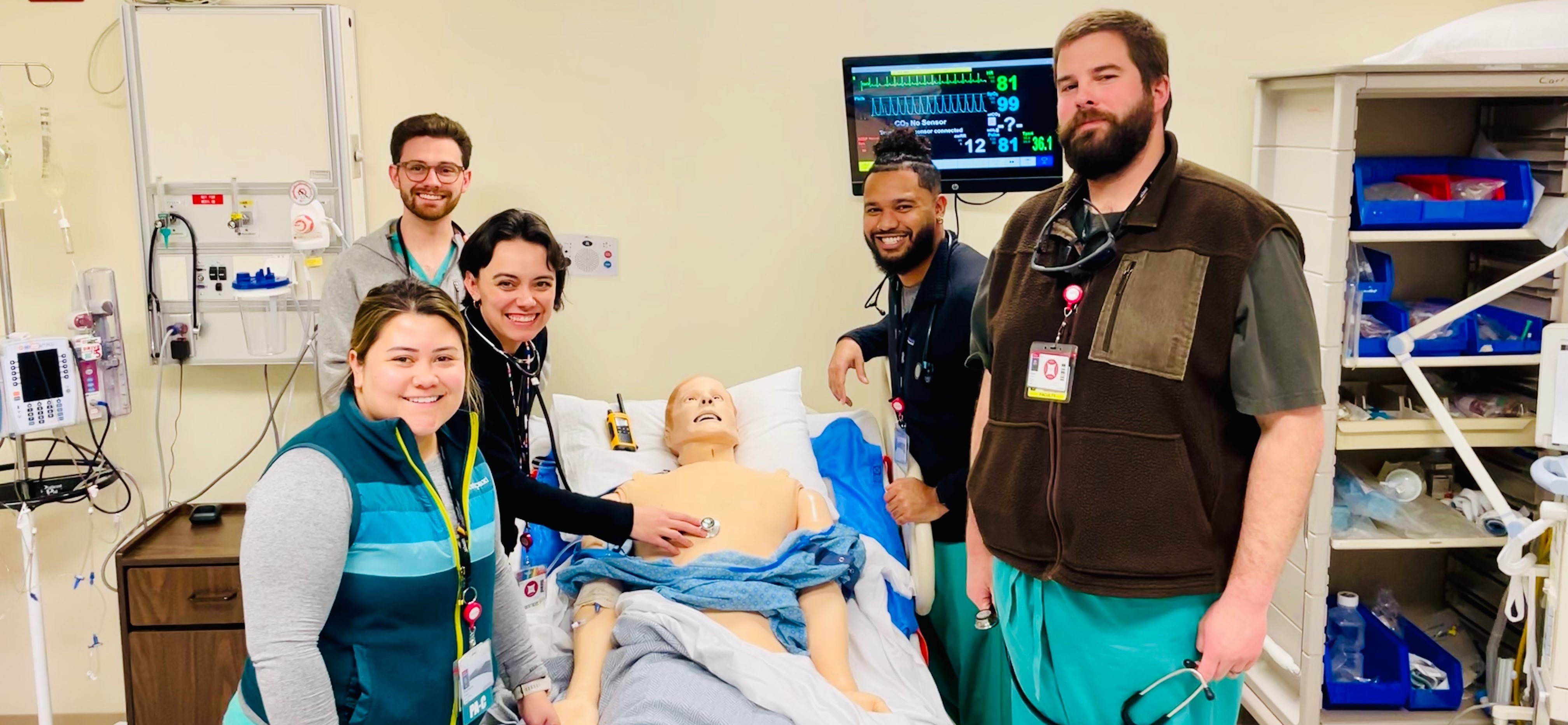 Photograph of 5 students in scrubs in a medical patient room. On the table is a mannequin used for training. In the background are a series of medical machines and supplies.
