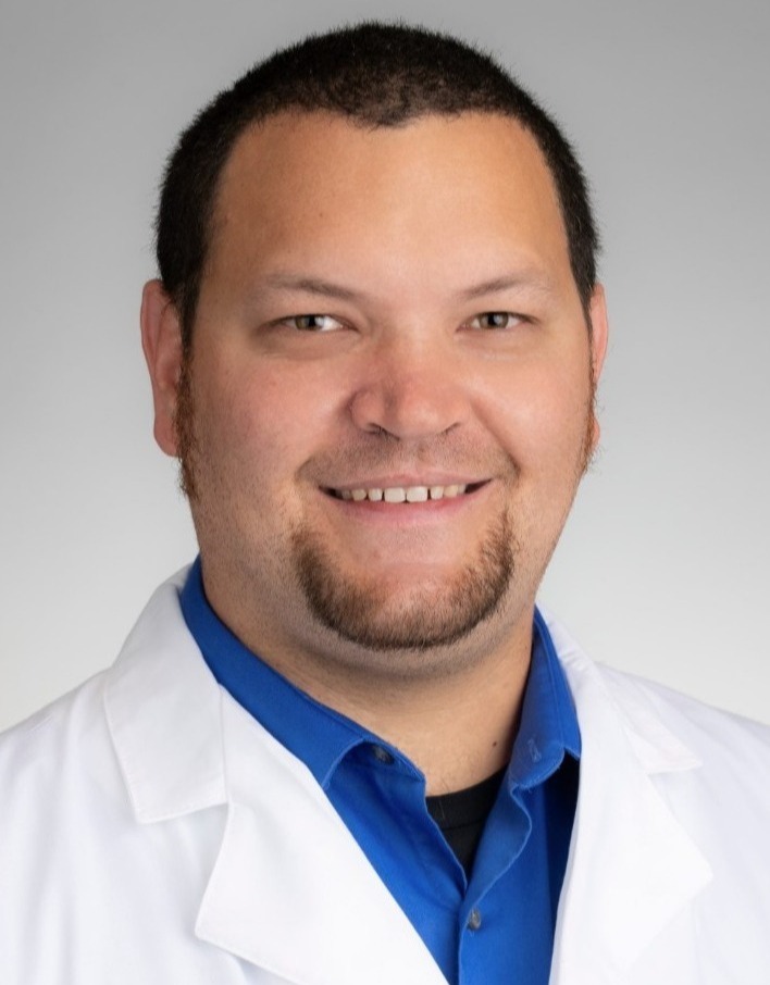 Headshot of man in collared shirt and white coat. Background is blank.