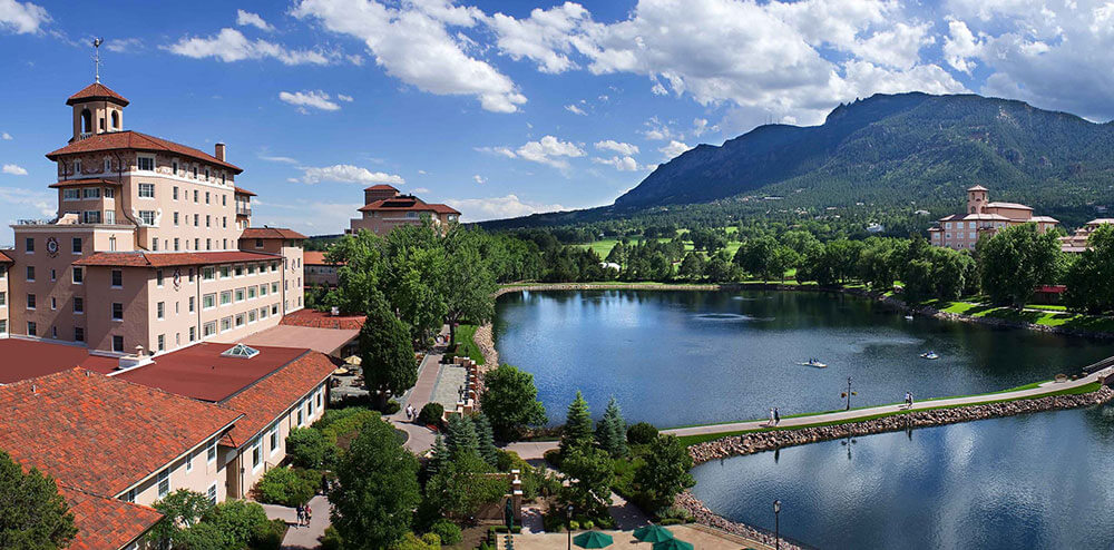 A scenic view of The Broadmoor resort in Colorado Springs, showing red-roofed buildings around a calm lake with a stone walkway, lush greenery, and Cheyenne Mountain rising in the background under a bright blue sky.