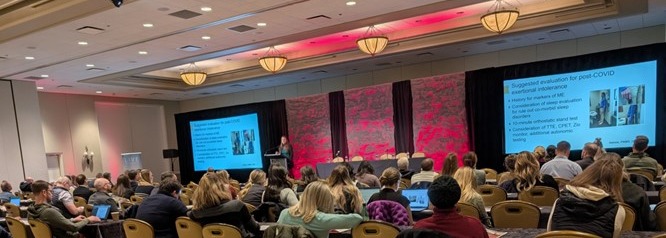 Audience seated in a conference room listening to a speaker presenting slides on post-COVID evaluation.