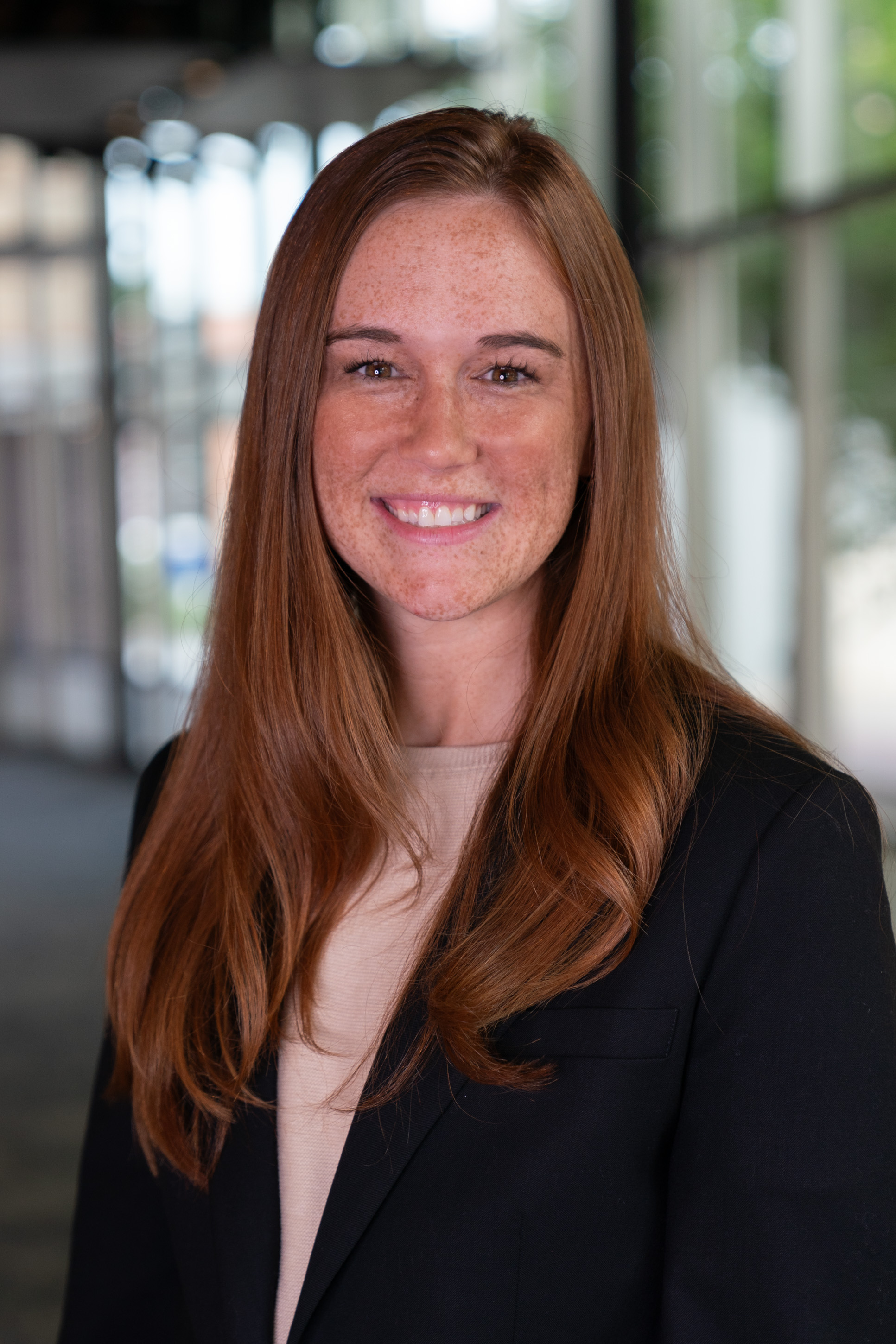 Professional headshot of woman in blazer. Background is a blurred hallway.