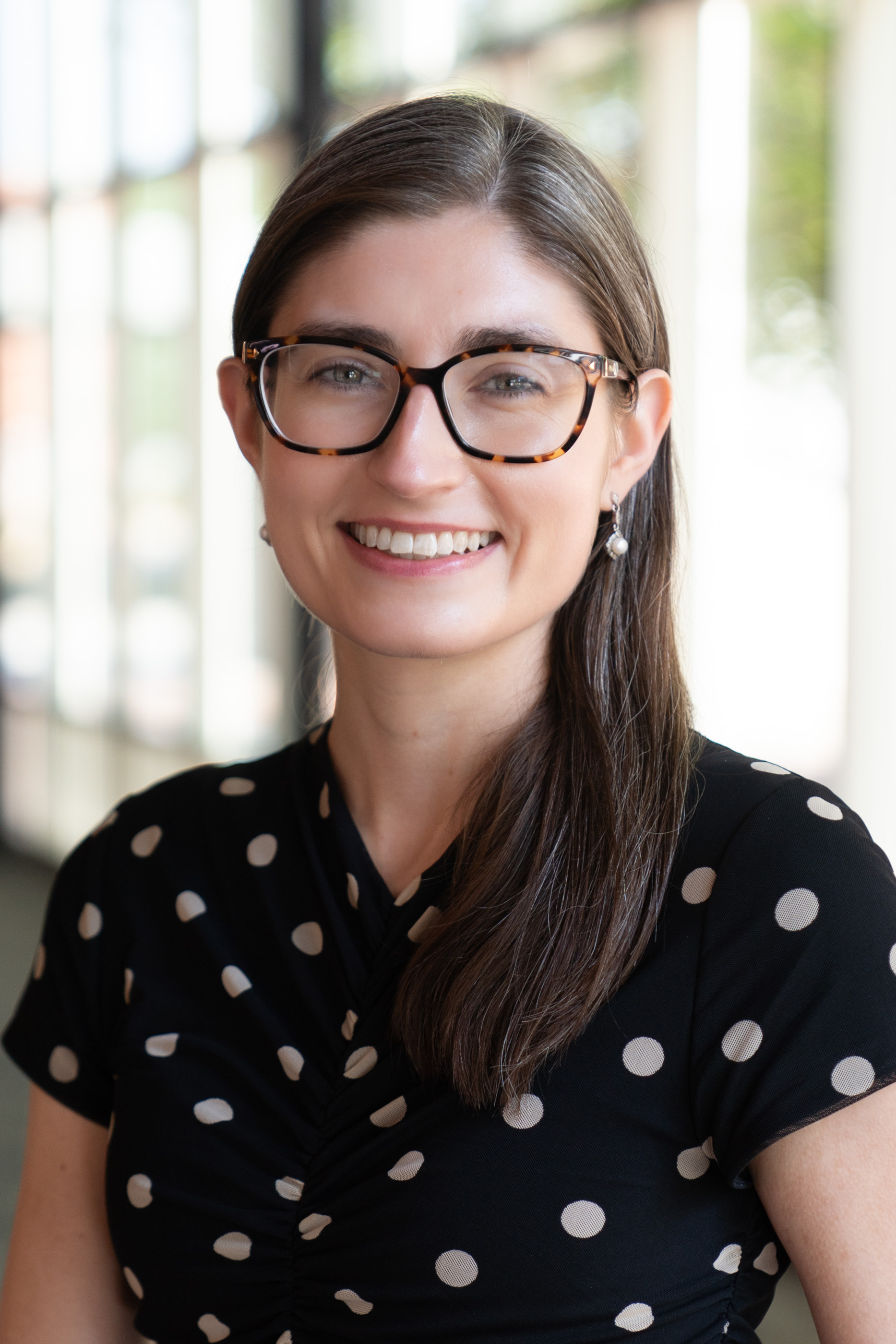Smiling woman with long brown hair, tortoiseshell glasses, pearl earrings, and a black polka-dot dress, photographed in a bright hallway.