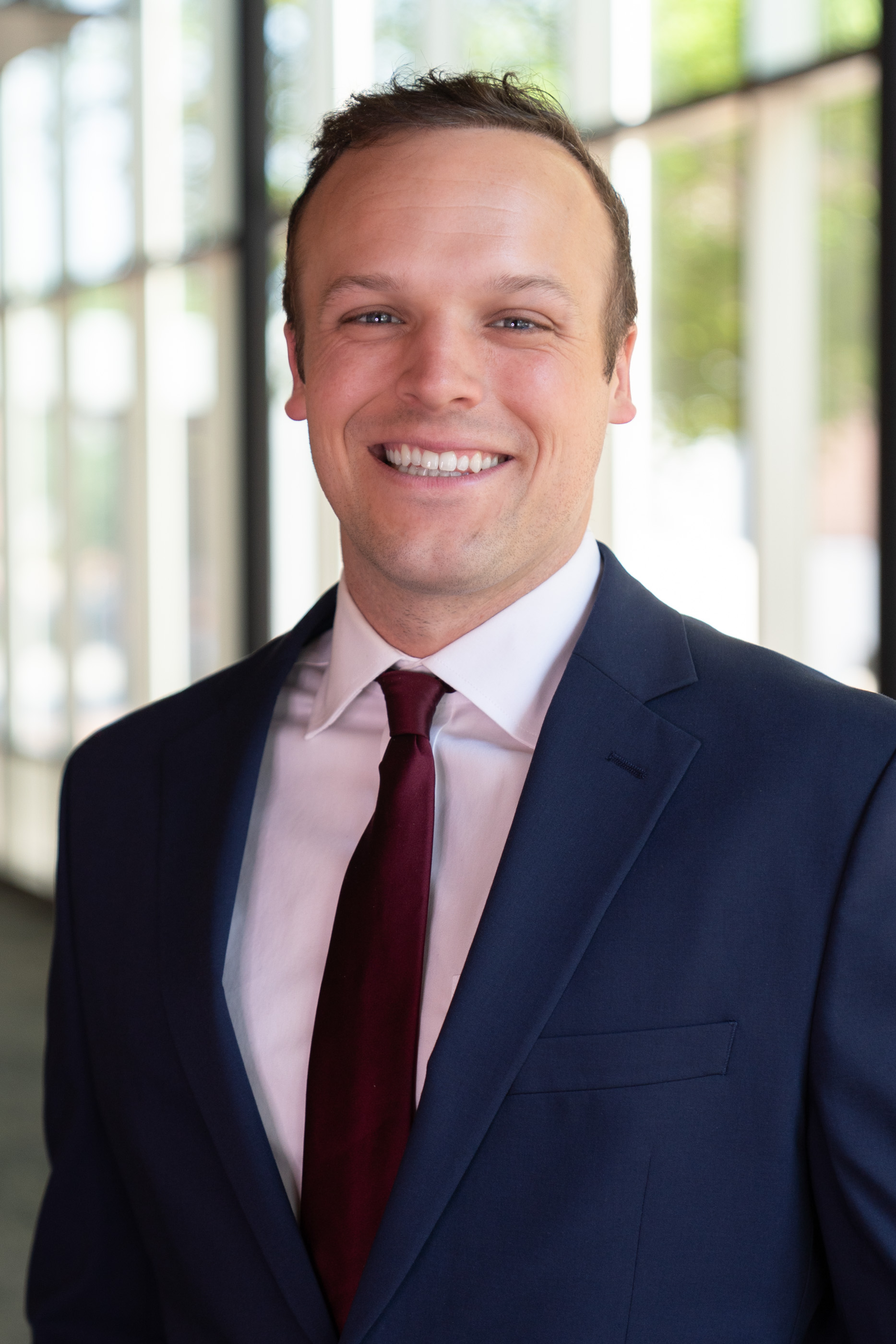 Smiling man wearing a navy suit, light dress shirt, and burgundy tie, photographed in a bright hallway.