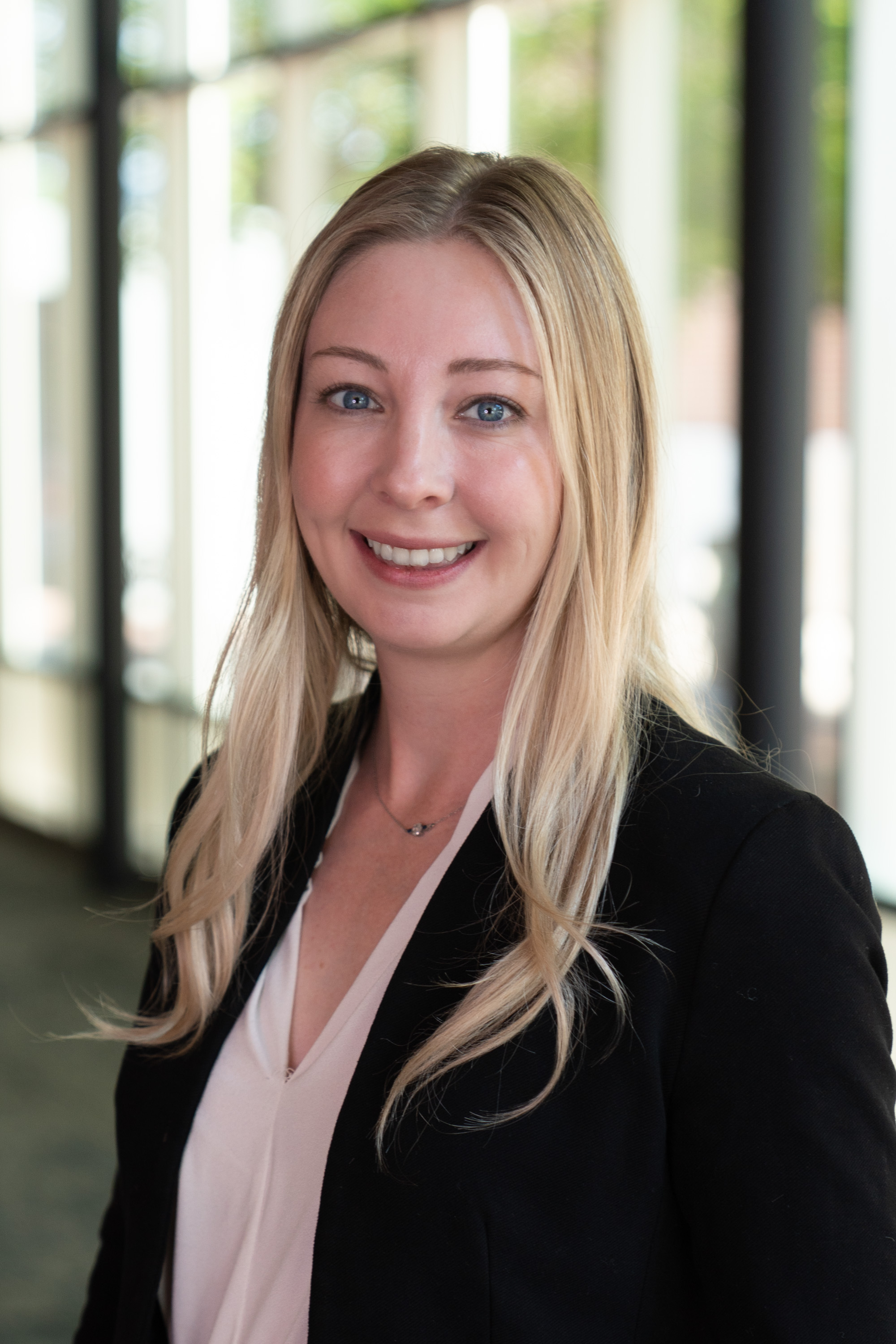 Smiling blonde woman wearing a black blazer over a pale blouse and a delicate necklace, photographed in a bright hallway.
