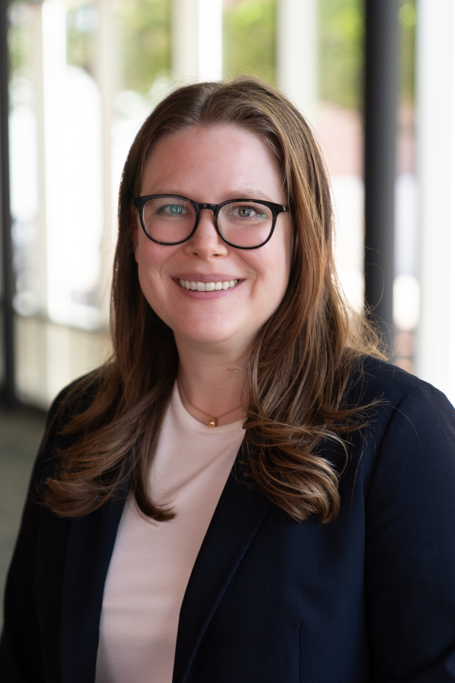 Smiling woman with long brown hair and black glasses wearing a navy blazer over a light top, photographed in a bright hallway.