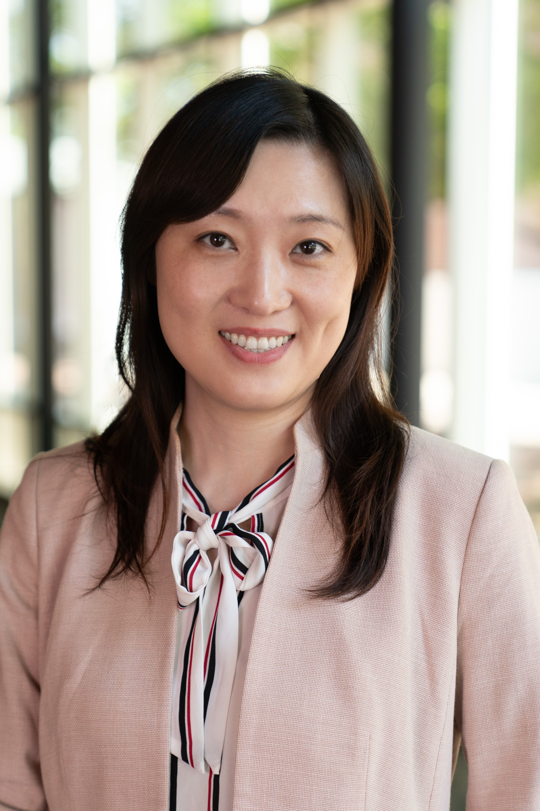 Smiling woman with dark hair wearing a pale pink blazer and a striped bow-tie blouse, photographed in a bright hallway.