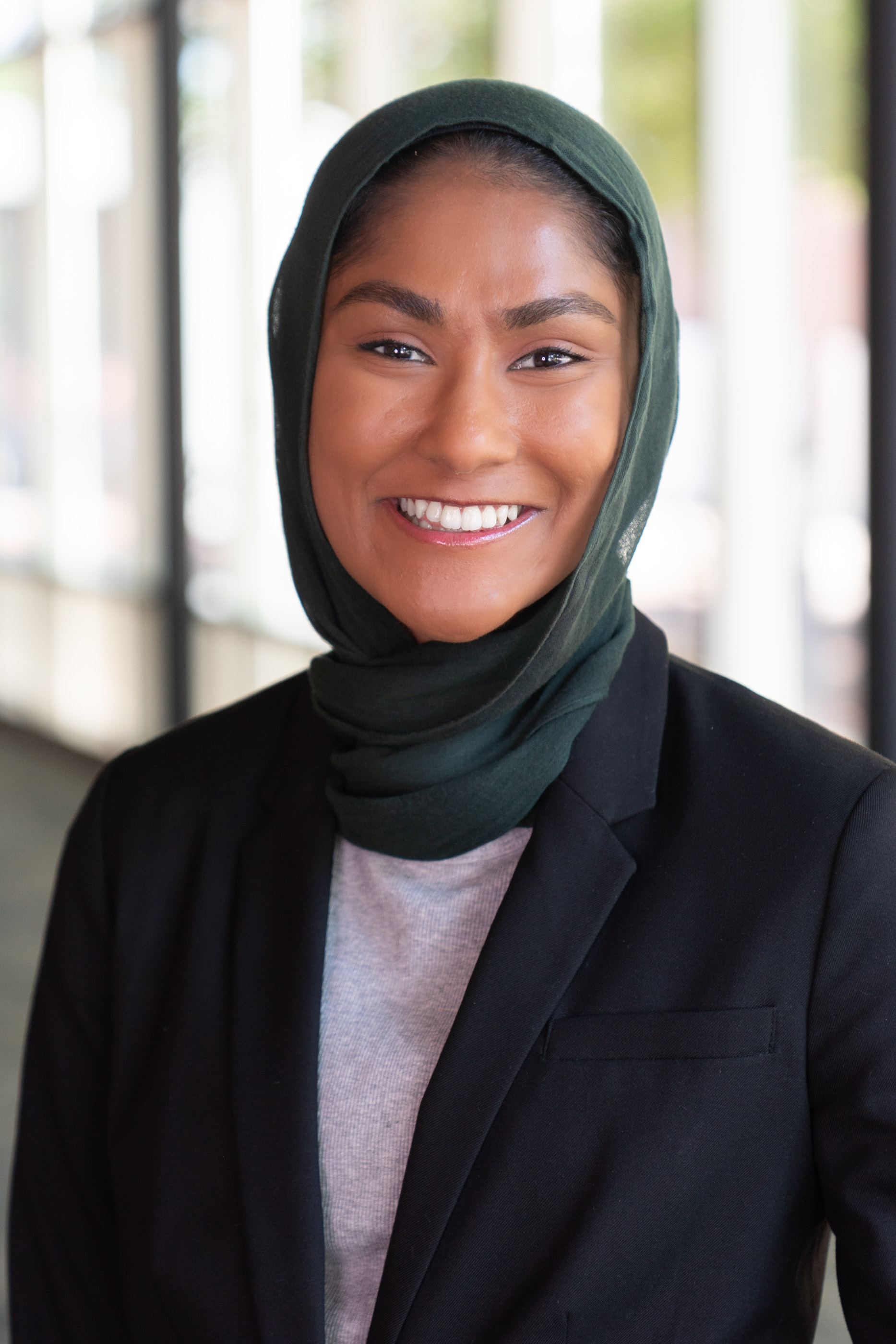 Smiling woman wearing a dark green hijab, black blazer, and gray top, photographed in a bright hallway.