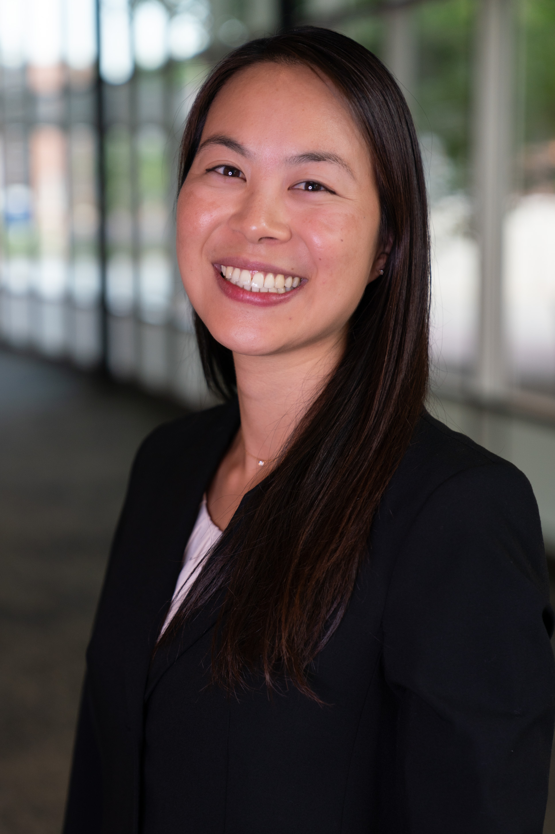 Professional headshot of women in blazer. Background is a blurred hallway.