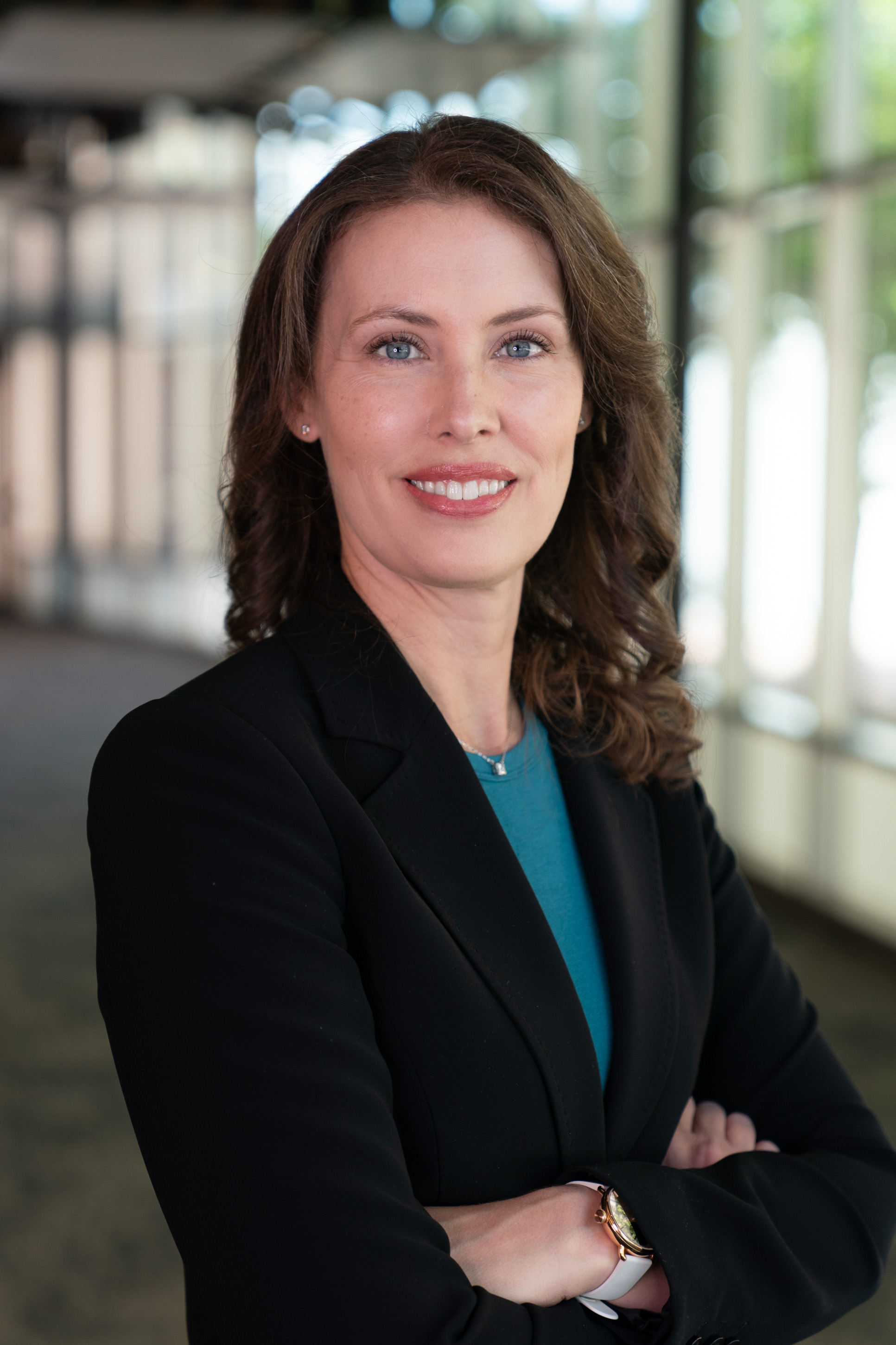 Professional headshot of woman in blazer. Background is blurry hallway.