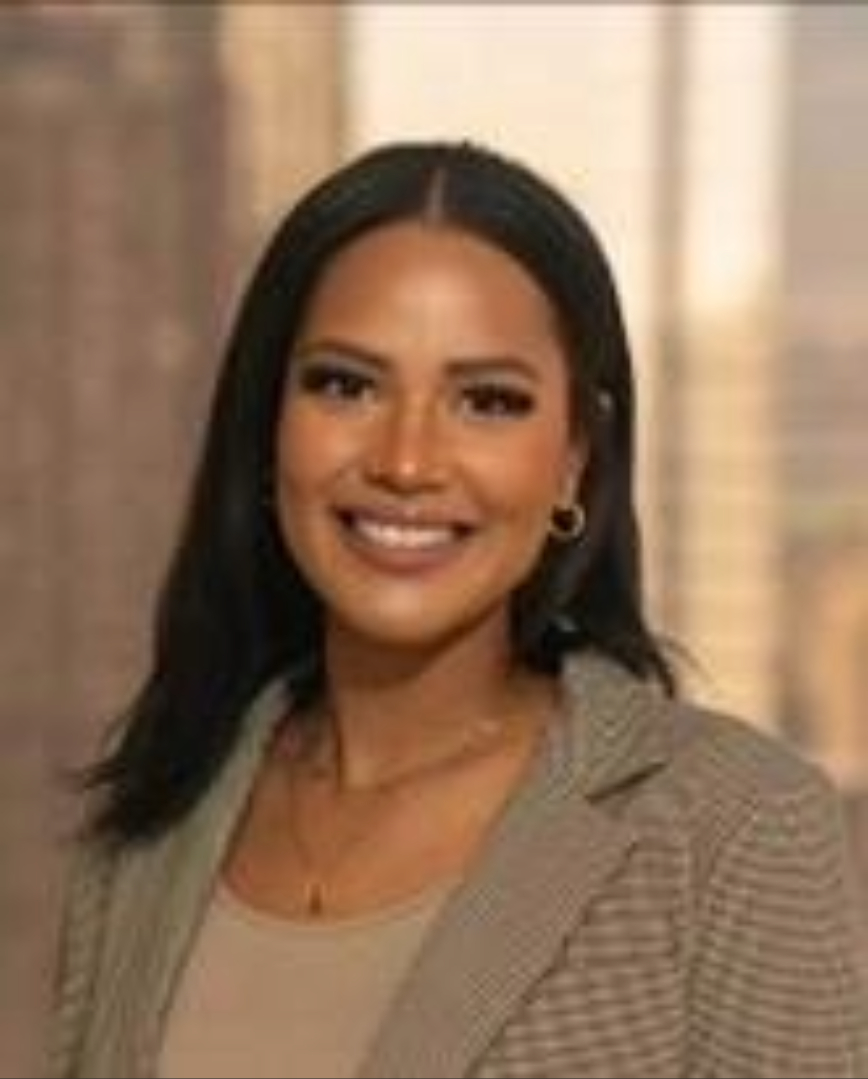 Professional photograph of a woman in a tan blazer and shirt. Background is a blurred city scape.