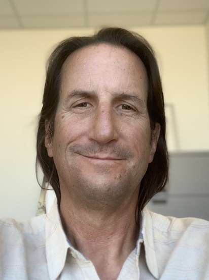 Headshot of a person with shoulder-length dark hair wearing a light button-down shirt, smiling gently at the camera in an office setting with a softly blurred background.
