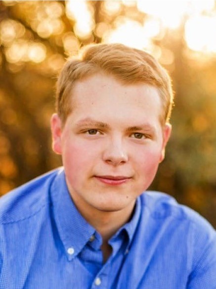 Head-and-shoulders outdoor portrait of a young person in a blue button-down shirt, looking at the camera with autumn trees and warm golden light softly blurred in the background.