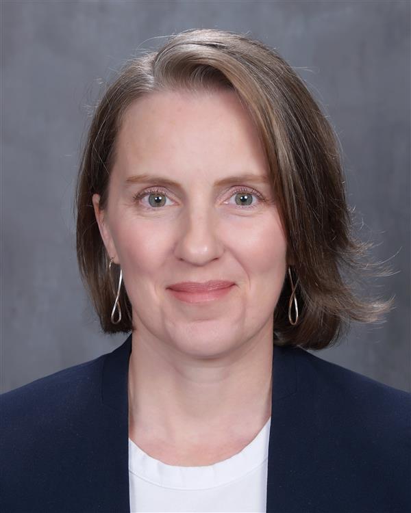 Professional headshot of a woman with short brown hair, wearing a navy blazer, white top, and silver drop earrings, smiling gently against a gray background.