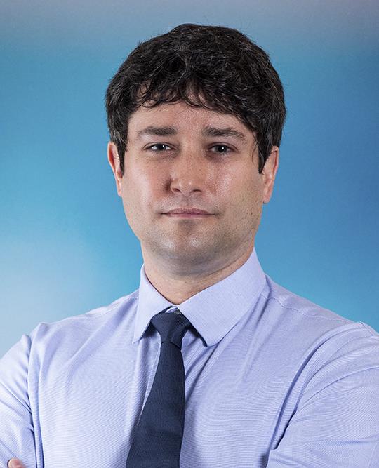 Professional headshot of man in shirt and tie. Background is plain blue backdrop