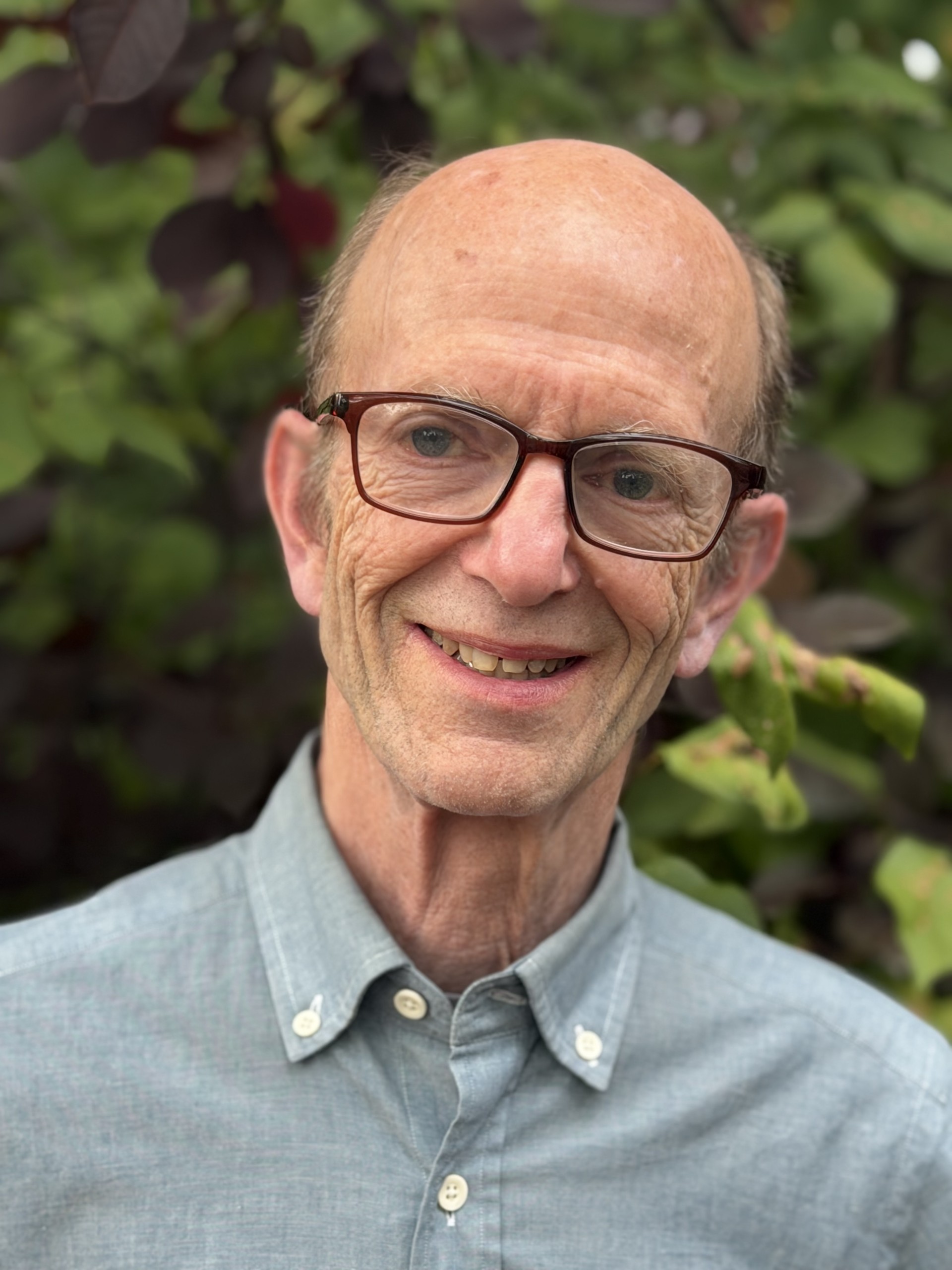 man wearing dark-framed glasses and a light blue button-down shirt smiles at the camera, with leafy greenery softly blurred in the background.