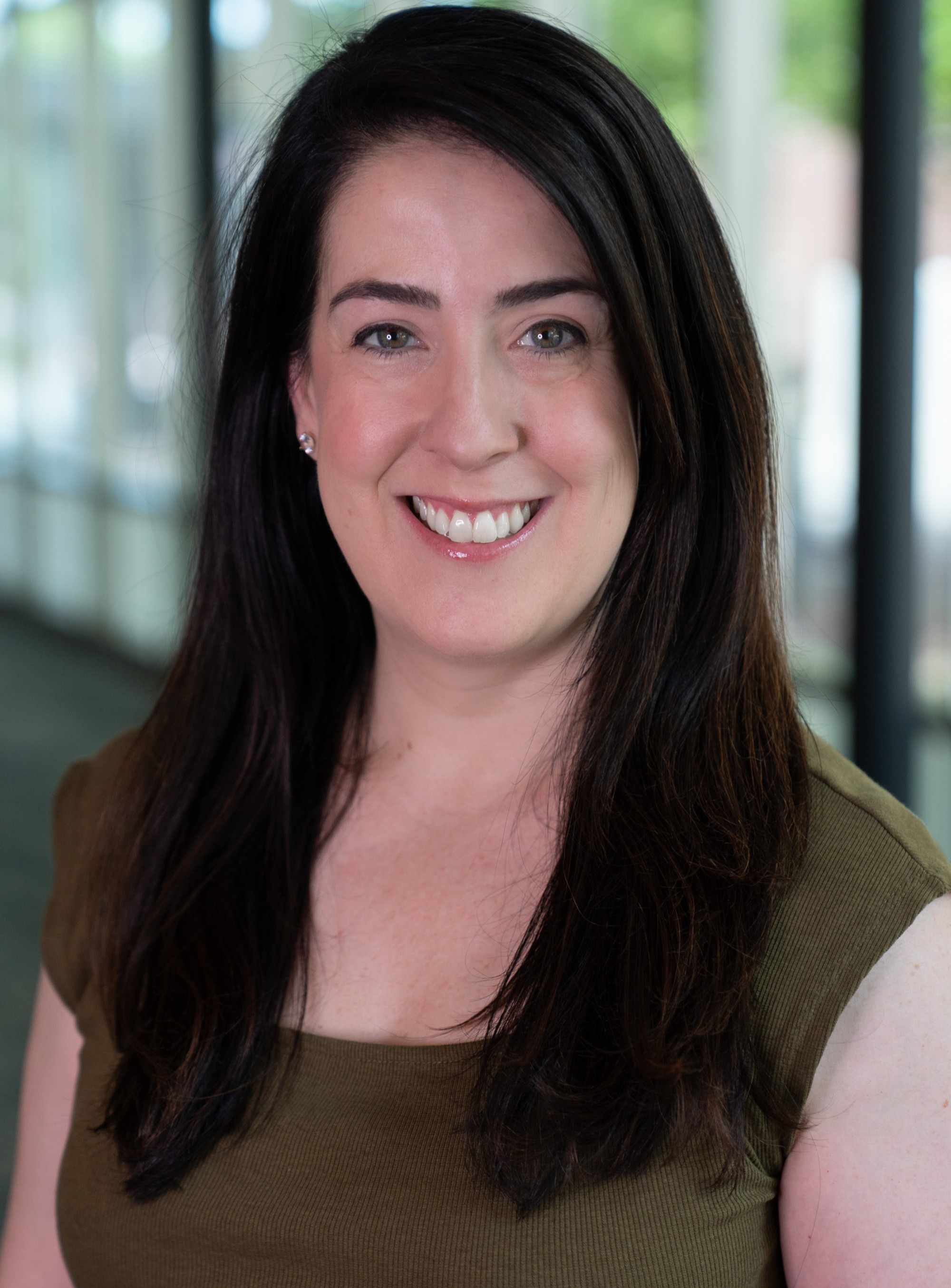 Professional headshot of a smiling person with long dark hair, wearing a sleeveless olive-green top, photographed indoors with a softly blurred office background.