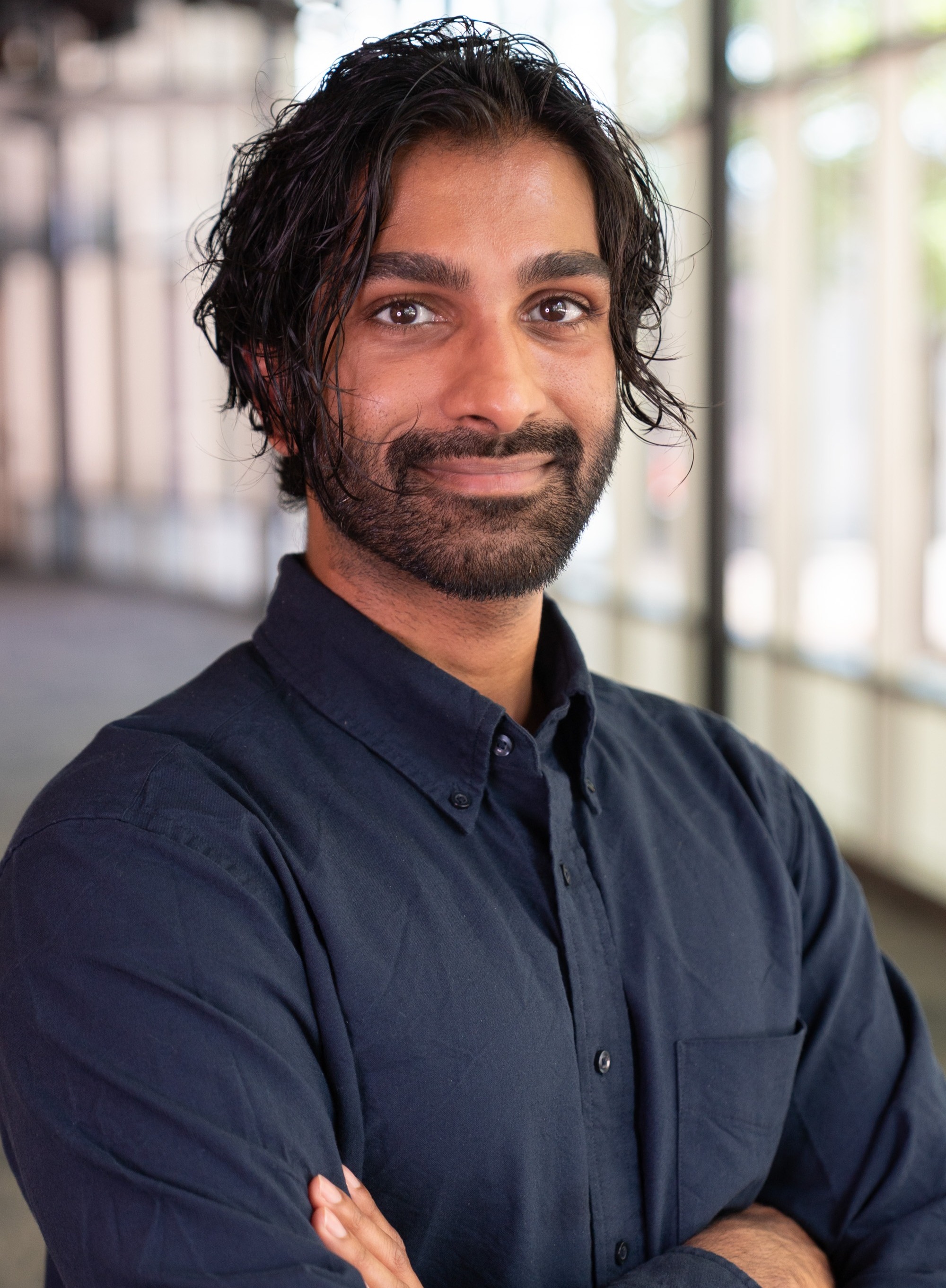 Professional headshot of a smiling person with medium-length dark hair and a beard, wearing a dark button-up shirt with arms crossed, photographed indoors with a blurred office background.