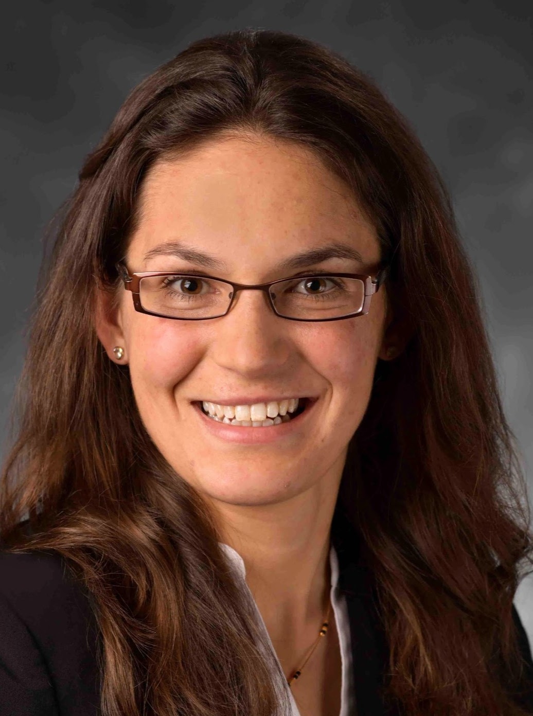 Professional headshot of a smiling person with long dark brown hair and glasses, wearing a black blazer over a white collared shirt, against a gray studio background.