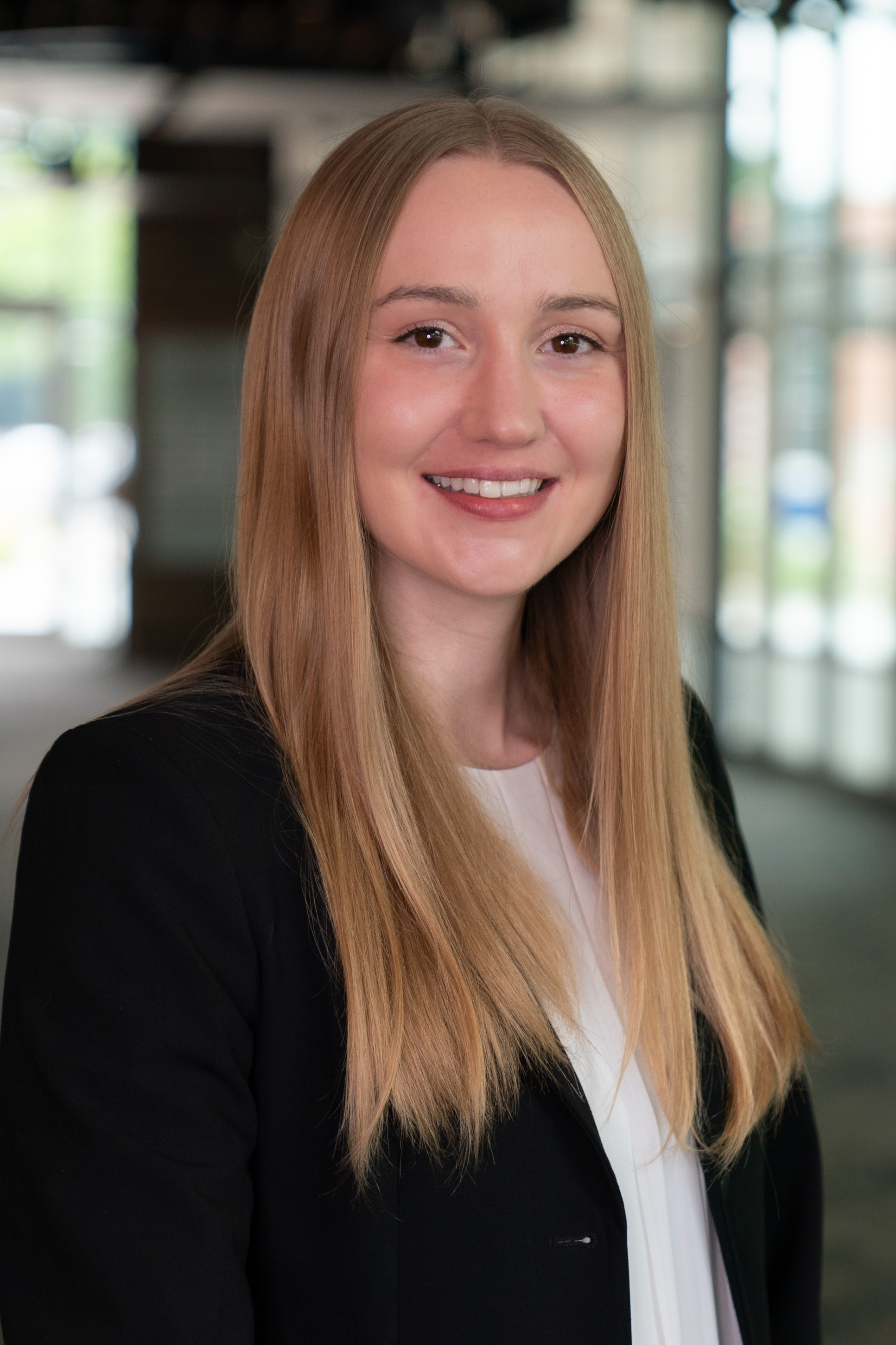 Professional headshot of woman in blazer. Background is a blurred hallway with large windows visible.