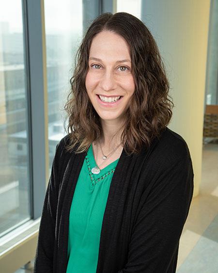 Professional headshot of women in sweater. The background is a blurred hallway, with large windows visible.