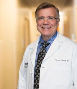 Professional headshot of man in white coat, shirt and tie underneath. Background is a blurred hallway.