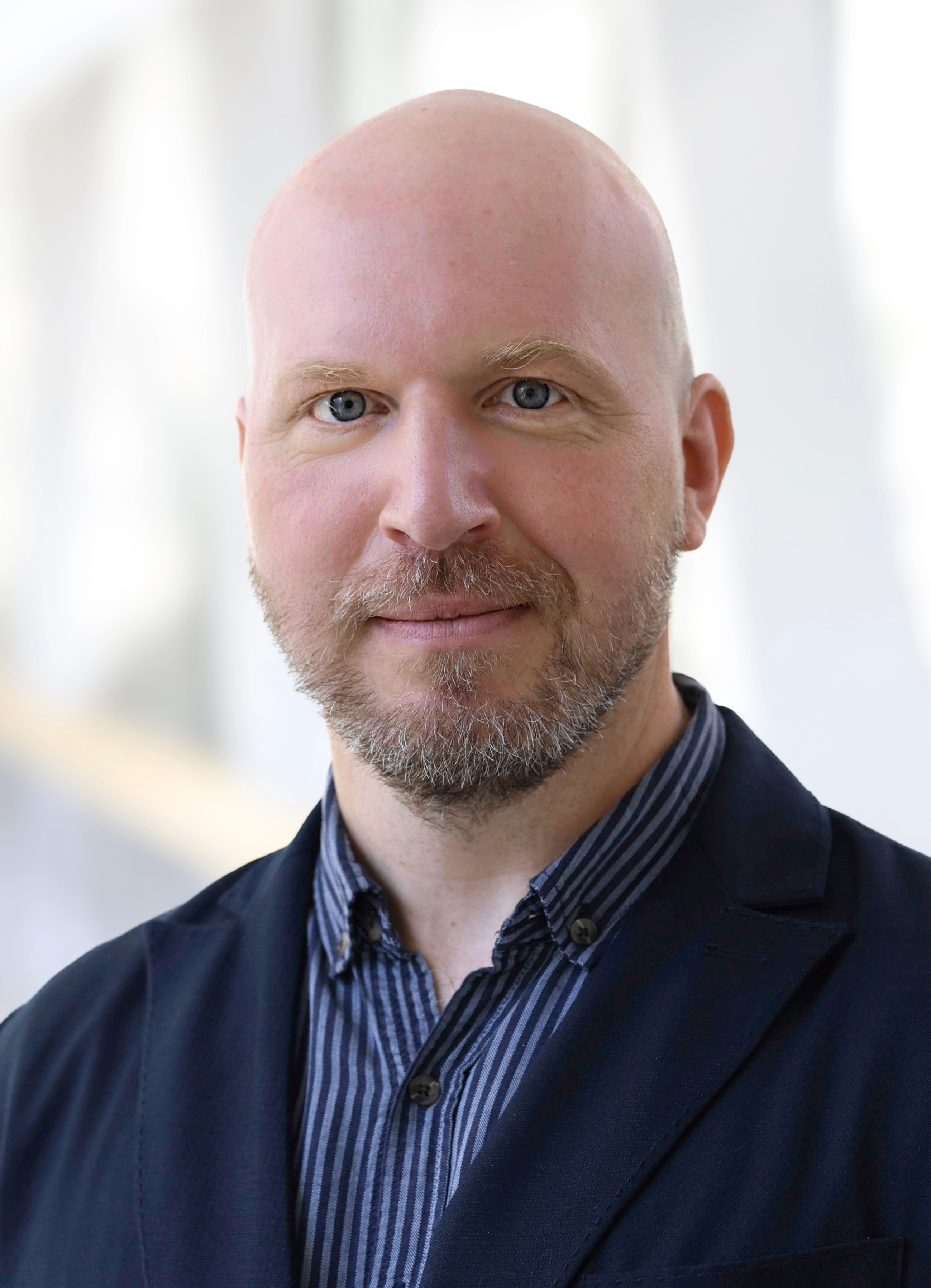 Professional headshot of a man with a short salt-and-pepper beard, wearing a dark navy blazer over a blue striped button-down shirt, against a softly blurred light background.