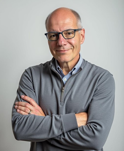 Professional portrait of a man with glasses and short gray hair, wearing a gray quarter-zip sweater over a collared shirt, standing with his arms crossed against a light gray background.