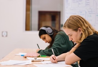 Two students sitting at a table writing. One student has earphones.
