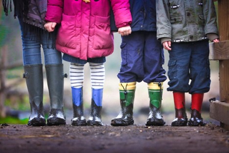 The legs only of 4 children in rainboots standing outside on wood.
