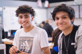 Two adolescent boys sitting in a classroom looking at the camera smiling.